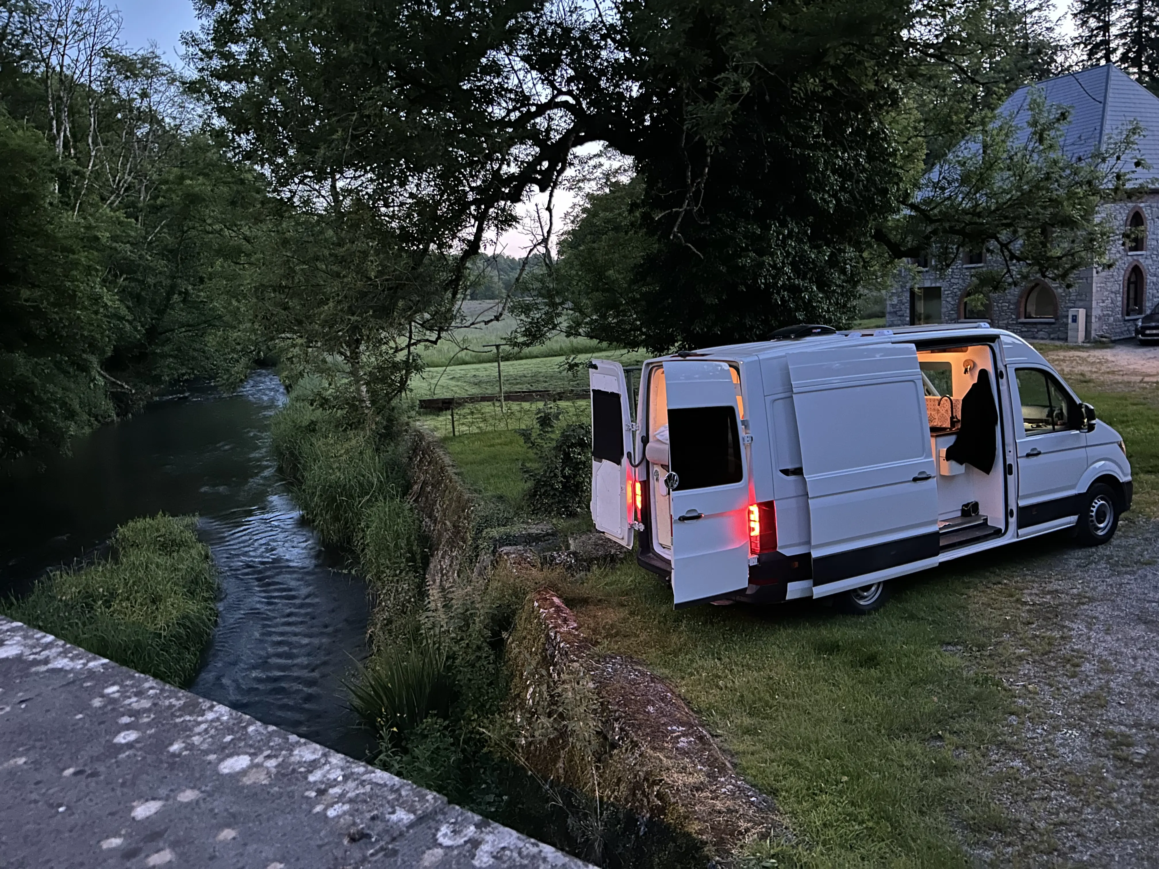 Camper van Granada Beach naast een riviertje bij valavond, achterdeuren open met een gloed van de achterlichten